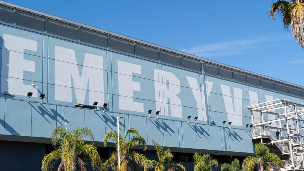 A wide shot of a building's grey facade says "EMERYVILLE." On the bottom and top right corner are palm trees.