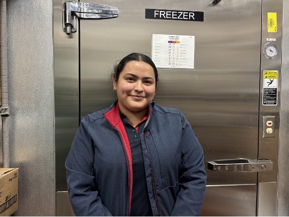 A woman with blue jacket stands in front of a commercial freezer.