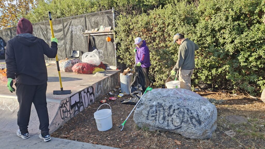 Three people with brooms, buckets, trash pickers and bags clean p a park, picking stuff up behind a graffitied rock and next to a graffittied concrete platform.