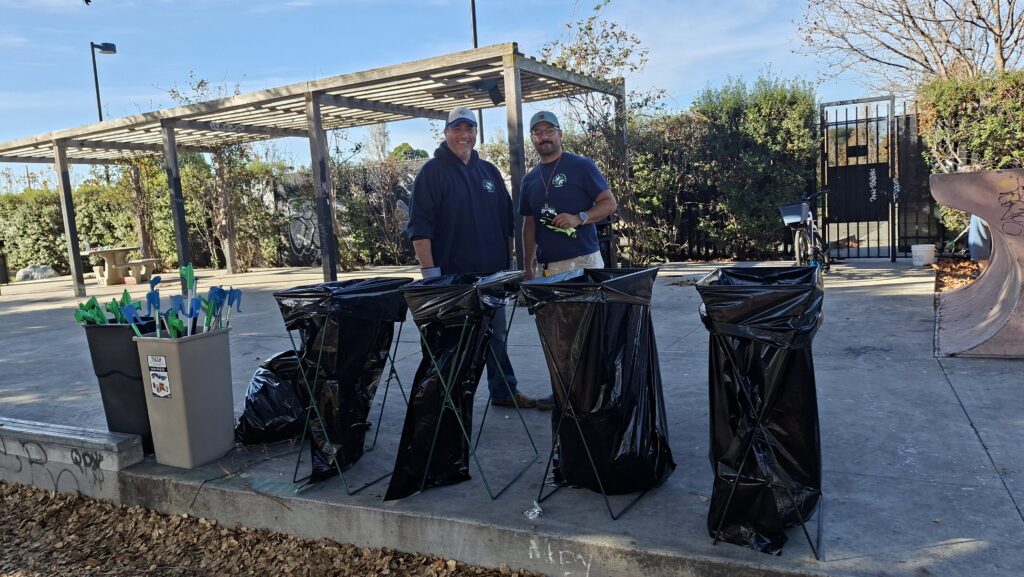 Two mean in baseball caps and navy tops stand before four open trash bags and two buckets of trash pickers on a concrete platform in a park.
