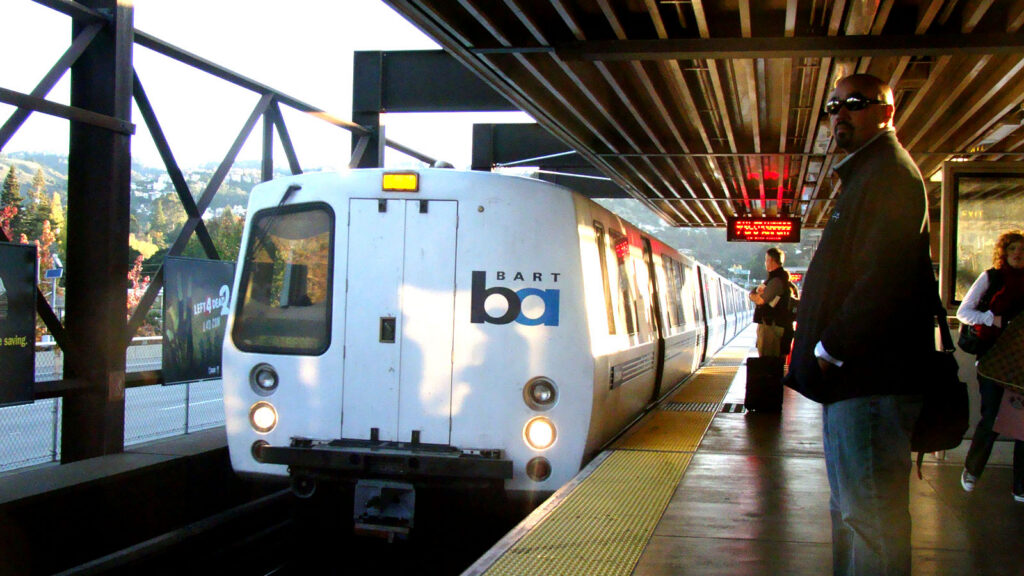 A BART train pulls into an outdoor station with people standing on the platform