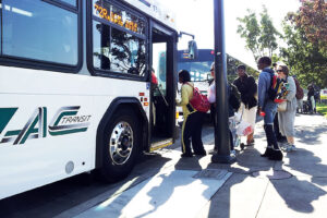 The right side of an AC Transit bus, with half the logo visible on the white side panel. ABout five people are lined up at a bus shelter, boarding through the front door.