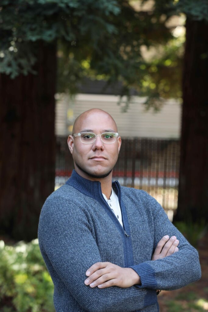 A portrait of Brandon Soublet wearing clear-framed glasses and a blue sweater, standing with his arms crossed in front of trees.