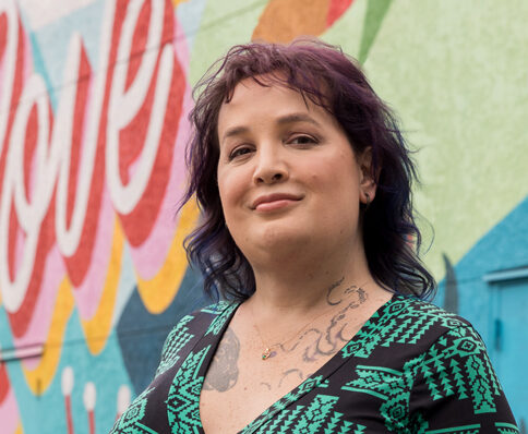 portrait of Emeryville City Council candifate Corry Frydlewicz smiling in front of a brightly painted wall mural that reads "Protect What You Love."