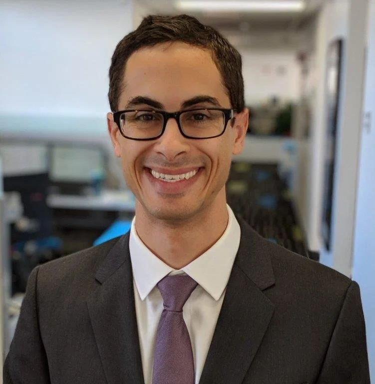 Headshot portrait of David Mourra smiling warmly in an office setting, wearing a dark suit, white dress shirt and purple tie.