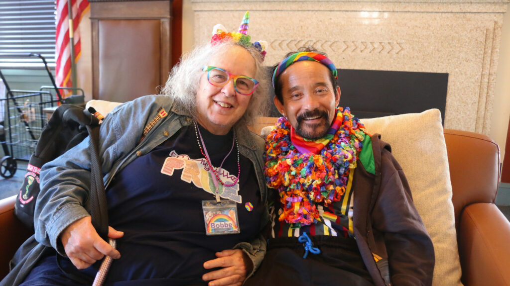 Two people sit together on a couch, posing to a camera. Both of them wear headwears, stickers and other rainbow-themed accessories.