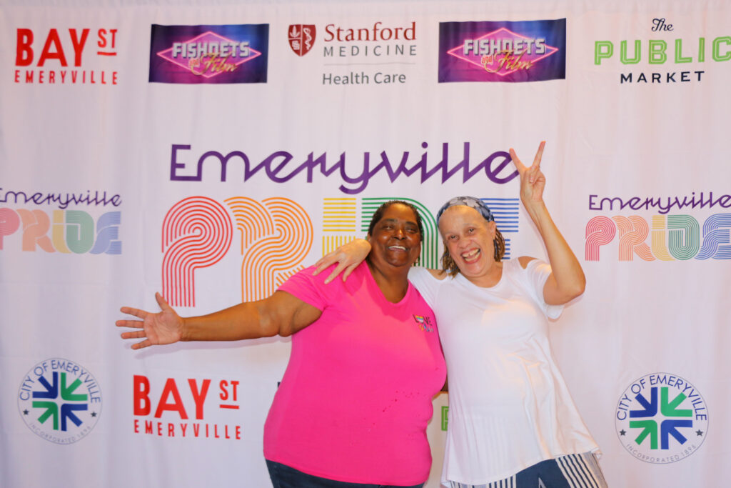 Two laughing people in t-shirts pose to a camera in front of a canvas.