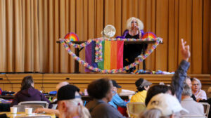 A dressed person sits by a table decorated with rainbow embellishments and flag on the stage of a hall. Under the stage are audiences, and one of them raises their hand.