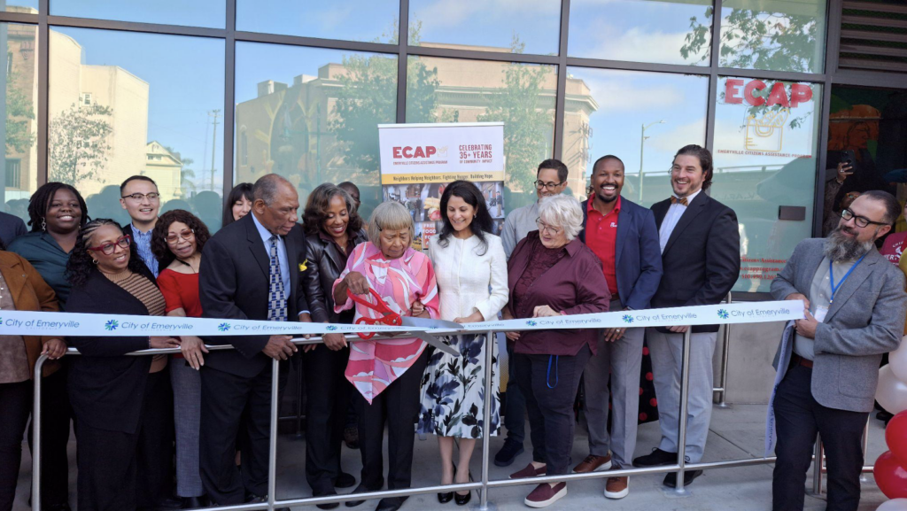 A old lady among group of people cut the ribbon in a ribbon-cutting ceremony in front of a residential builiding.