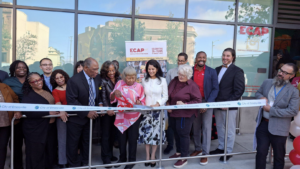 A old lady among group of people cut the ribbon in a ribbon-cutting ceremony in front of a residential builiding.