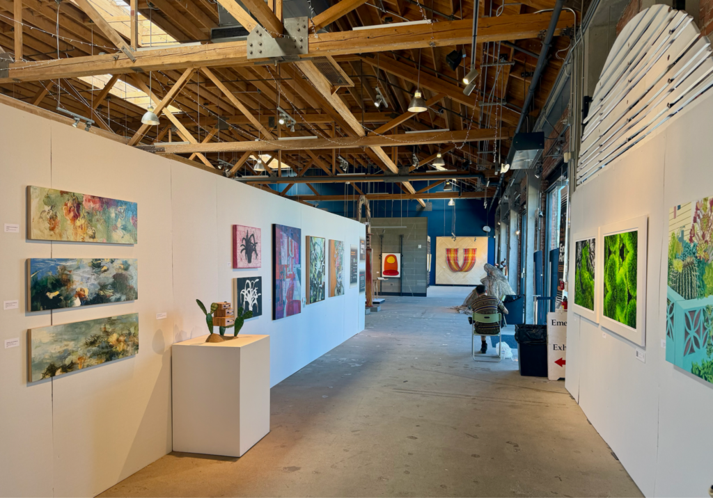In a large warehouse like room with a wood and metal industrial-type ceiling and cement floor, paintings hand on white walls.