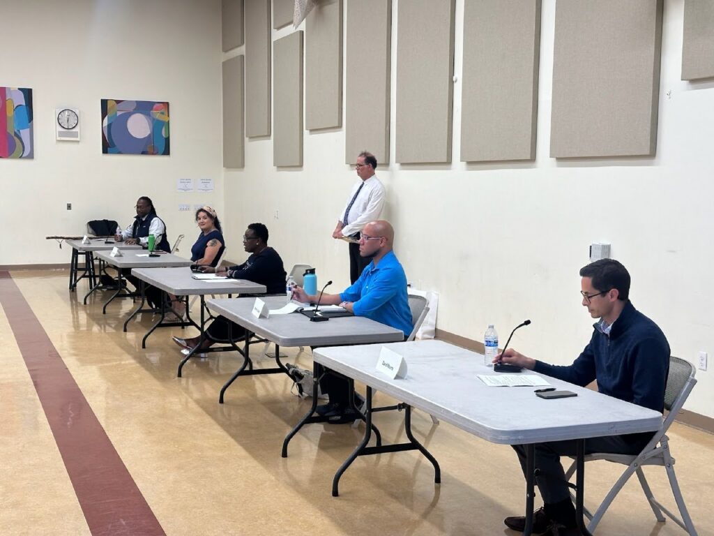 Five people sit in a line on folding chairs behind individual tables in a large room with a tan and brown linoleum floor.