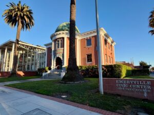 Shot of a building with a green dome, red siding and white pillars flanking the entrance, which is arched. A palm tree rises above the building in the middle of the frame and to the left is a glass sided extension of the domed building.