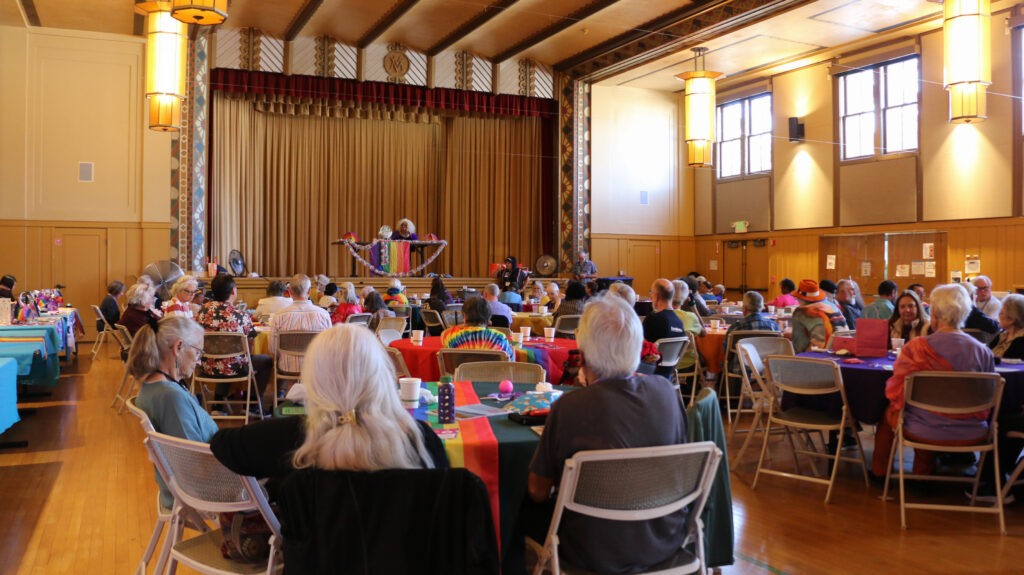 A hall packed with audiences. A person is speaking on the stage who sits by a table decorated with rainbow decorations